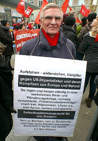 Demonstrant mit umgehängtem Schild: »Aufstehen - widerstehen, kämpfen gegen US-Imperialisten und deren Komplizen aus Europa und Nahost…«.